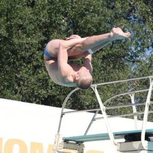 本·威利特(Ben Willett)在跳水时做了半空中翻转. 他的腿伸直，双臂放在膝盖后面. 背景是一个跳水板和绿色的树叶.